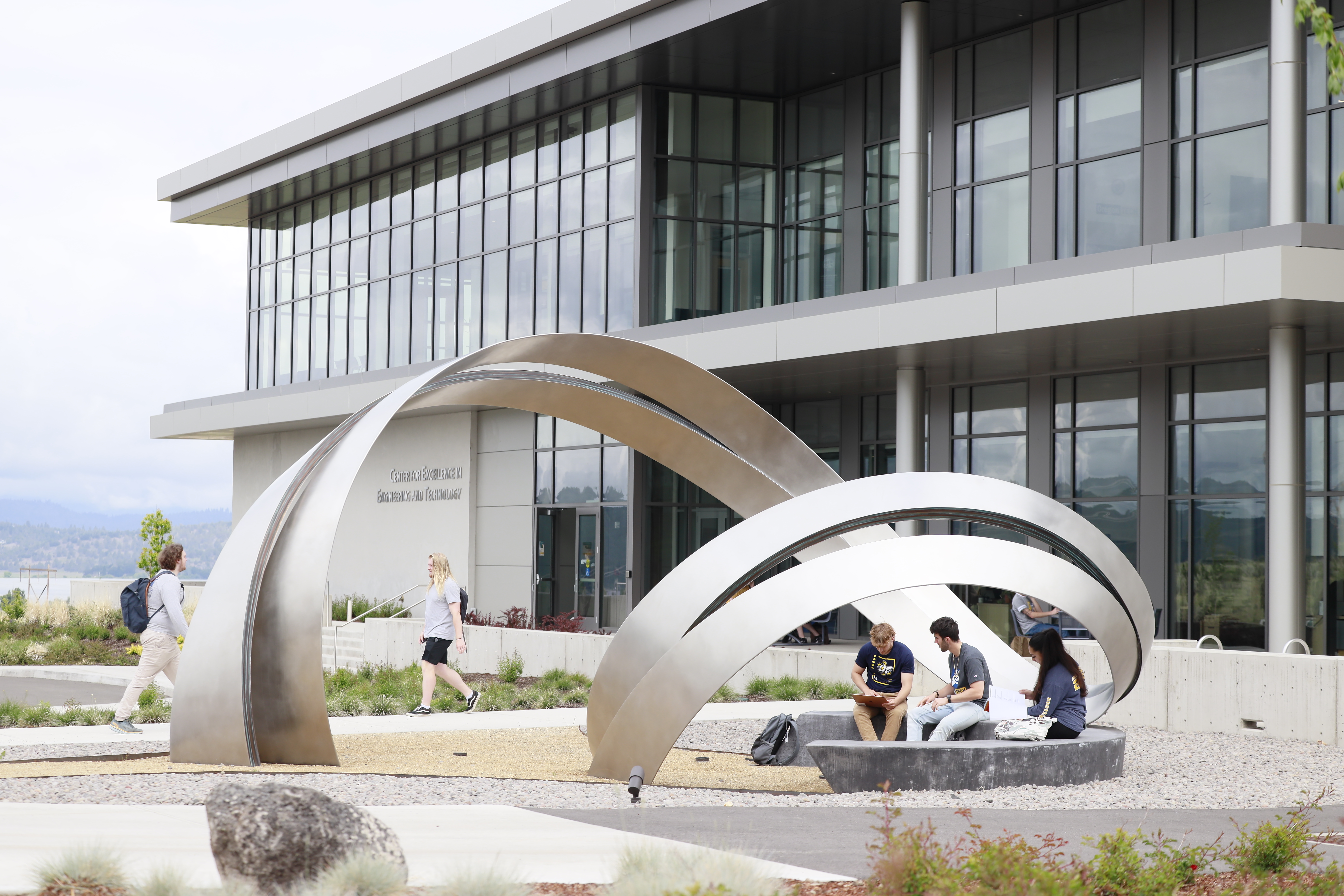 Students sitting under an abstract geometric sculpture near a large academic building